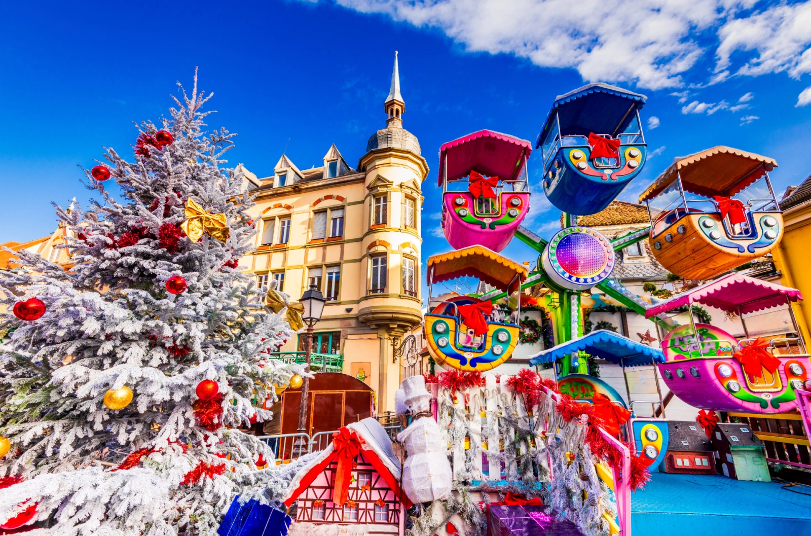 Snow-dusted Christmas market in Alsace with whimsical carnival rides, historic architecture, and a festive white tree under bright blue skies